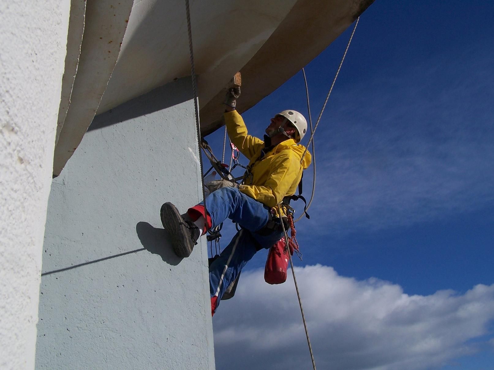 Un grimpeur en veste jaune descend en rappel une paroi grise sur fond de ciel bleu