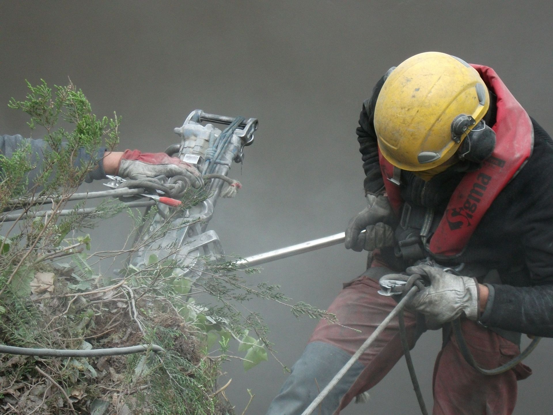 Un ouvrier portant un casque jaune utilise une corde au milieu des broussailles et de la poussière.