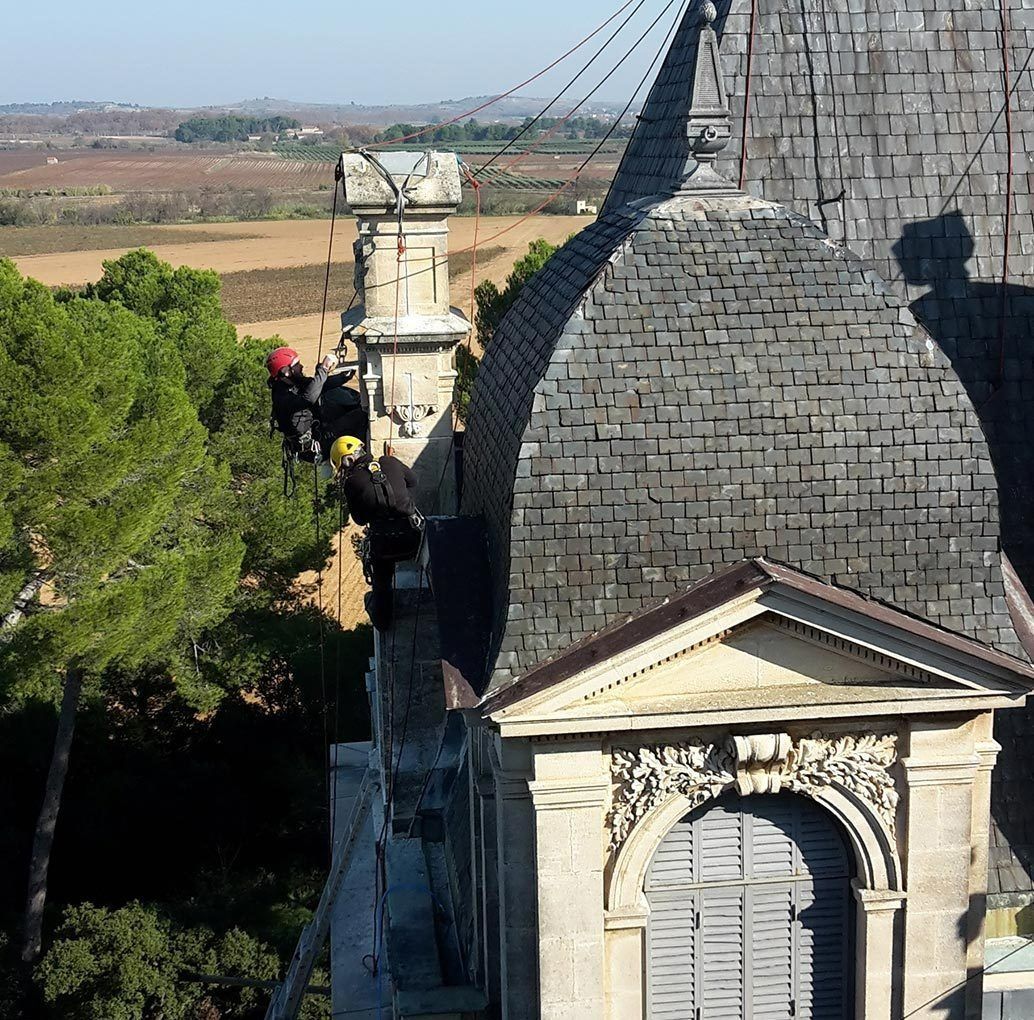 Bâtiment historique en pierre avec un toit d'ardoise pentu, et un cordiste travaillant sur un ouvrage bétonné