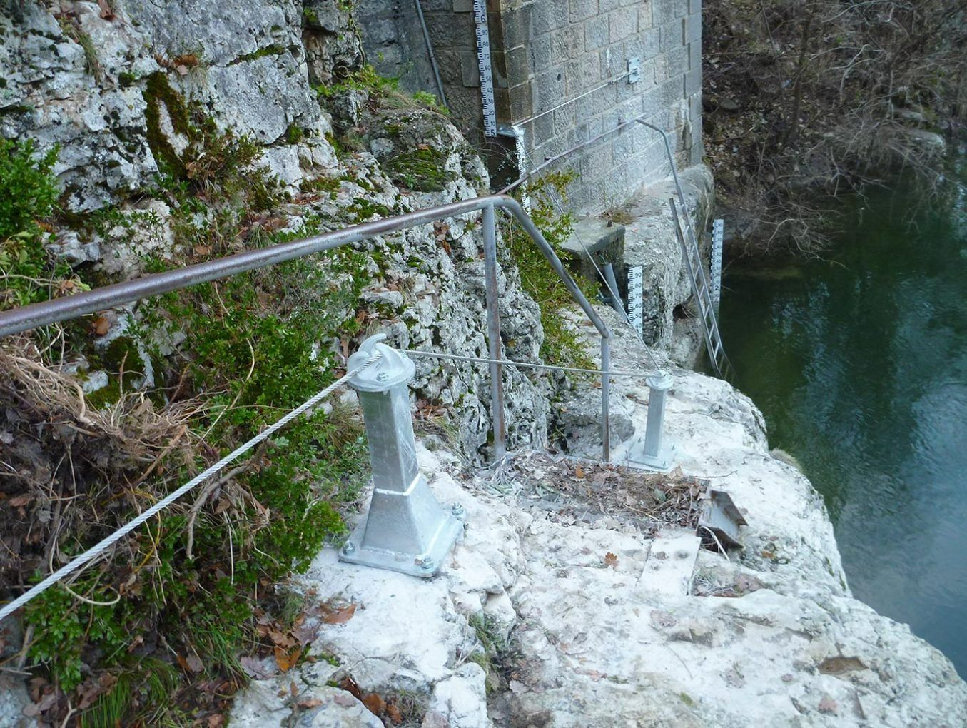 Sentier escarpé en pierre avec rambarde métallique longeant une falaise surplombant des eaux vertes