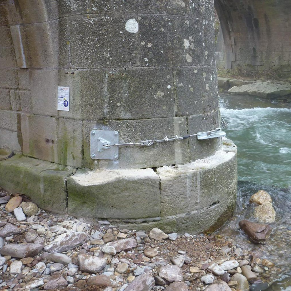 Pile de pont en béton au bord d'une rivière, avec une rive rocheuse et une ligne de vie métallique