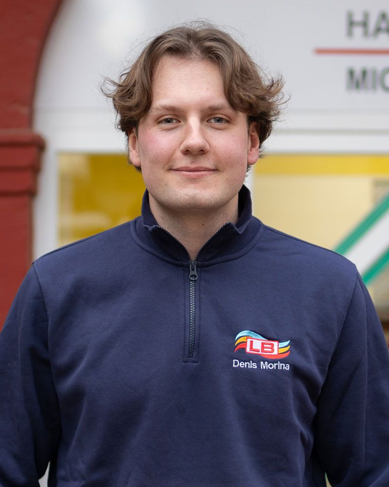 Ein junger Mann mit lockigem Haar lächelt in die Kamera und trägt ein kariertes Hemd.