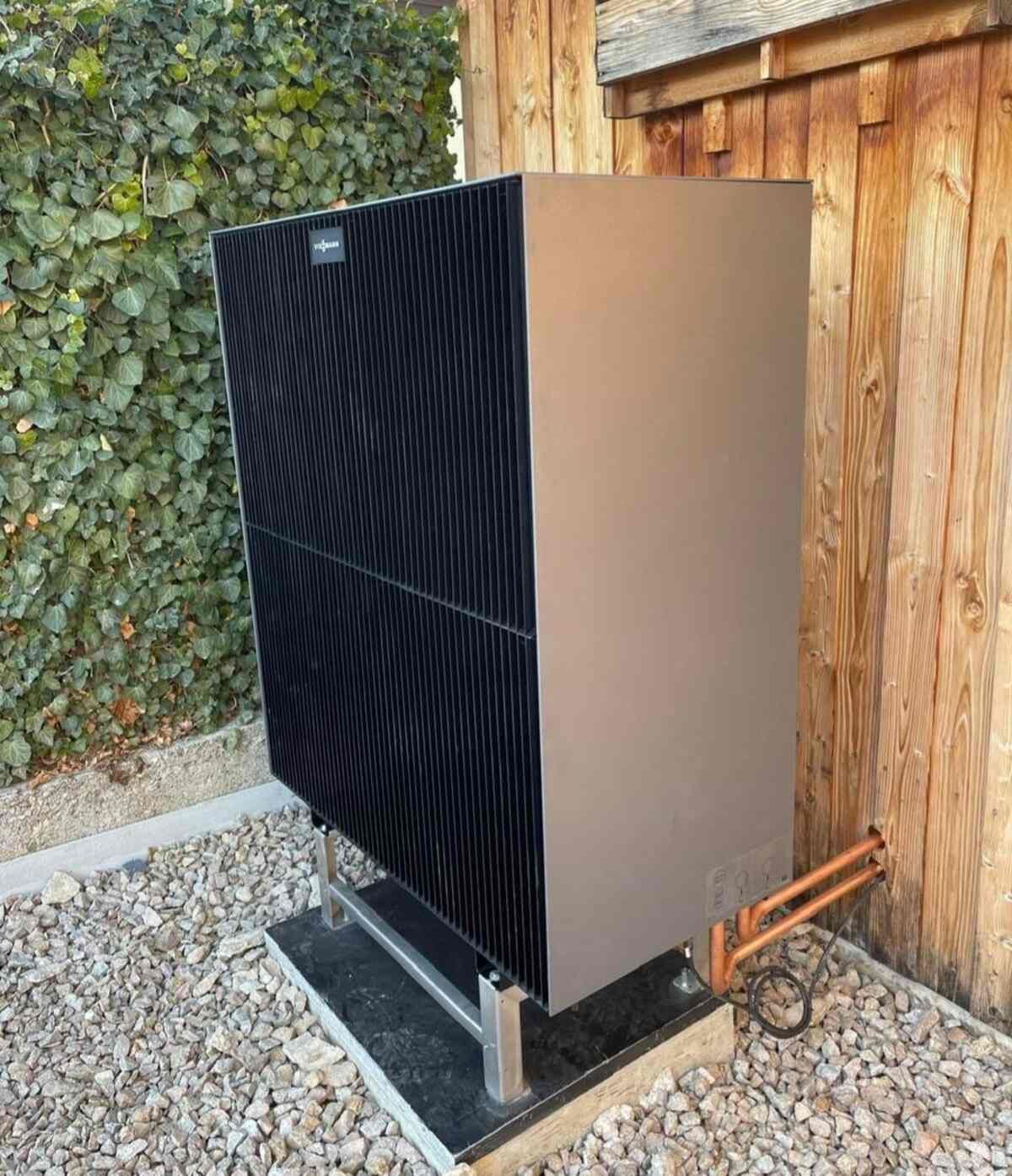 Eine Wärmepumpe steht auf einem Kieshaufen neben einer Holzmauer.