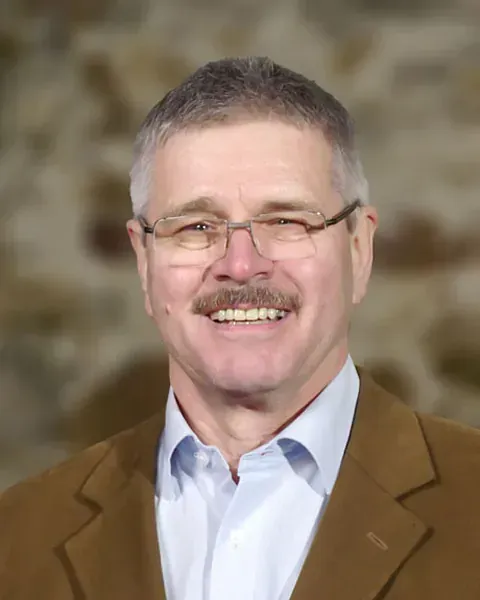 Ein Mann mit Brille und Schnurrbart lächelt vor einer Steinmauer.