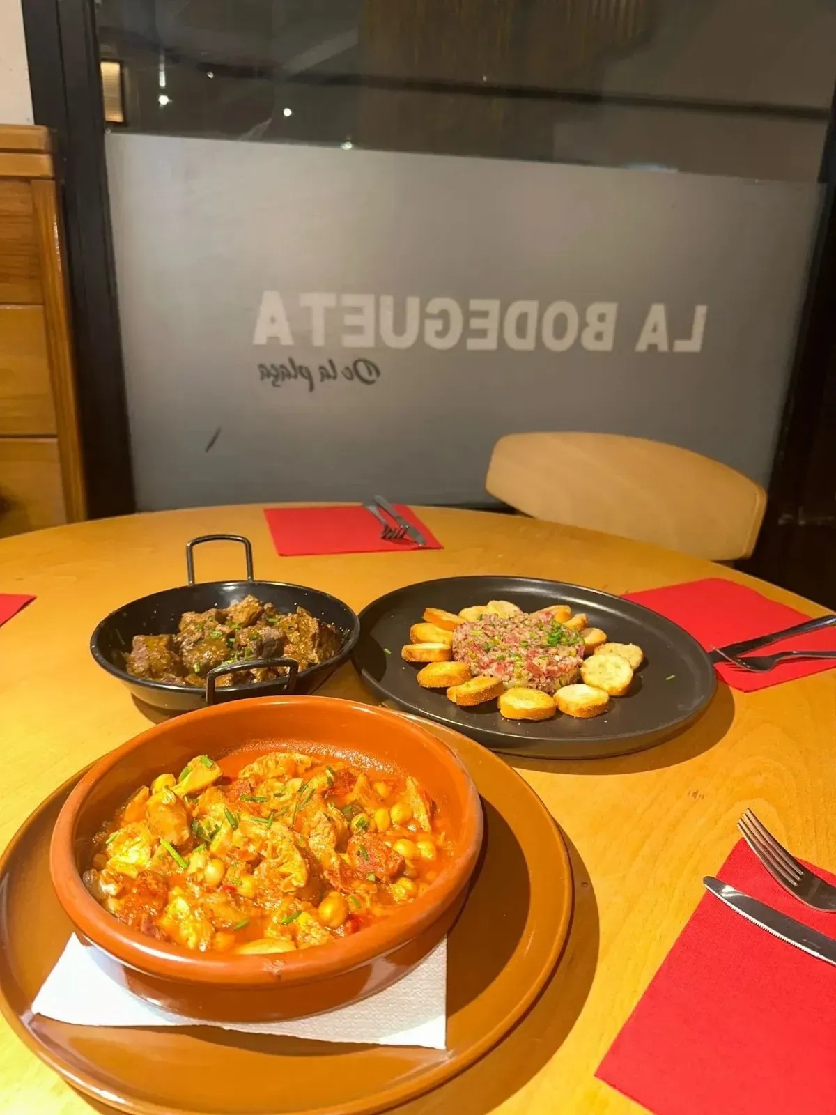 Mesa con tapas españolas: estofado, patatas bravas, berenjenas con el logotipo del restaurante de fondo.