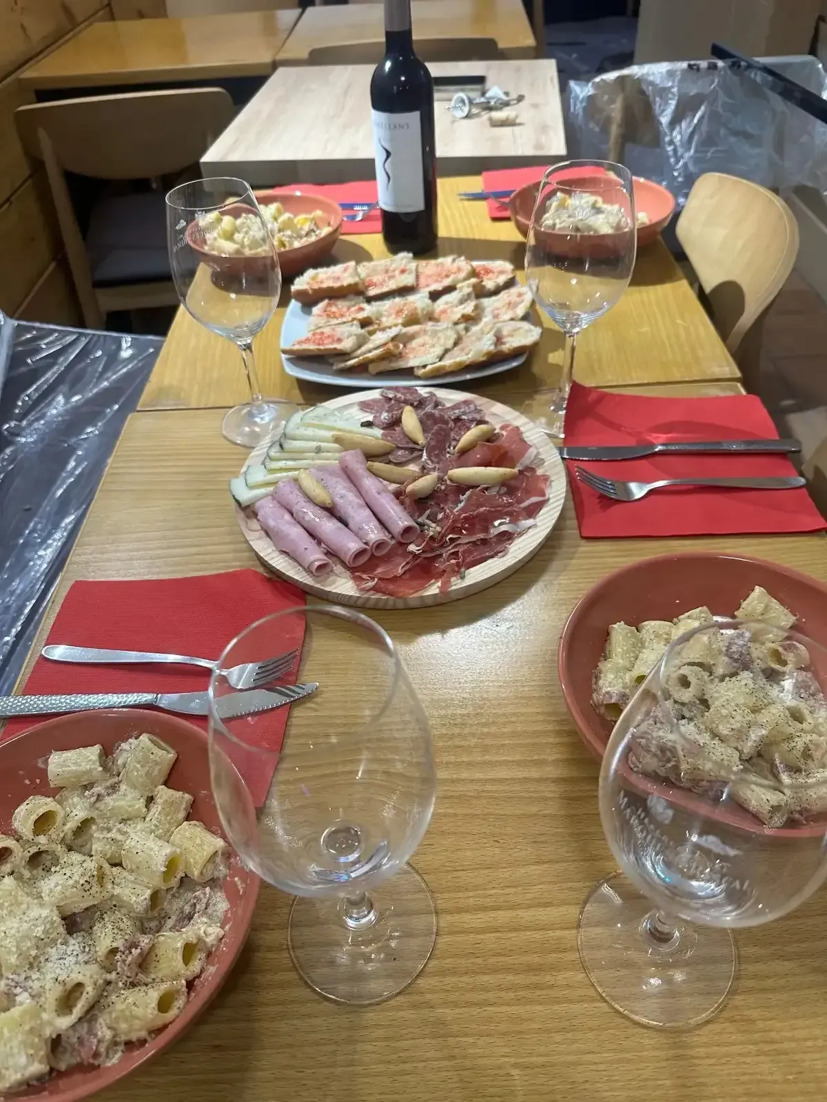 Mesa preparada para comer: pasta, embutidos, pan, copas y una botella de vino. Mesa de madera con servilletas rojas.