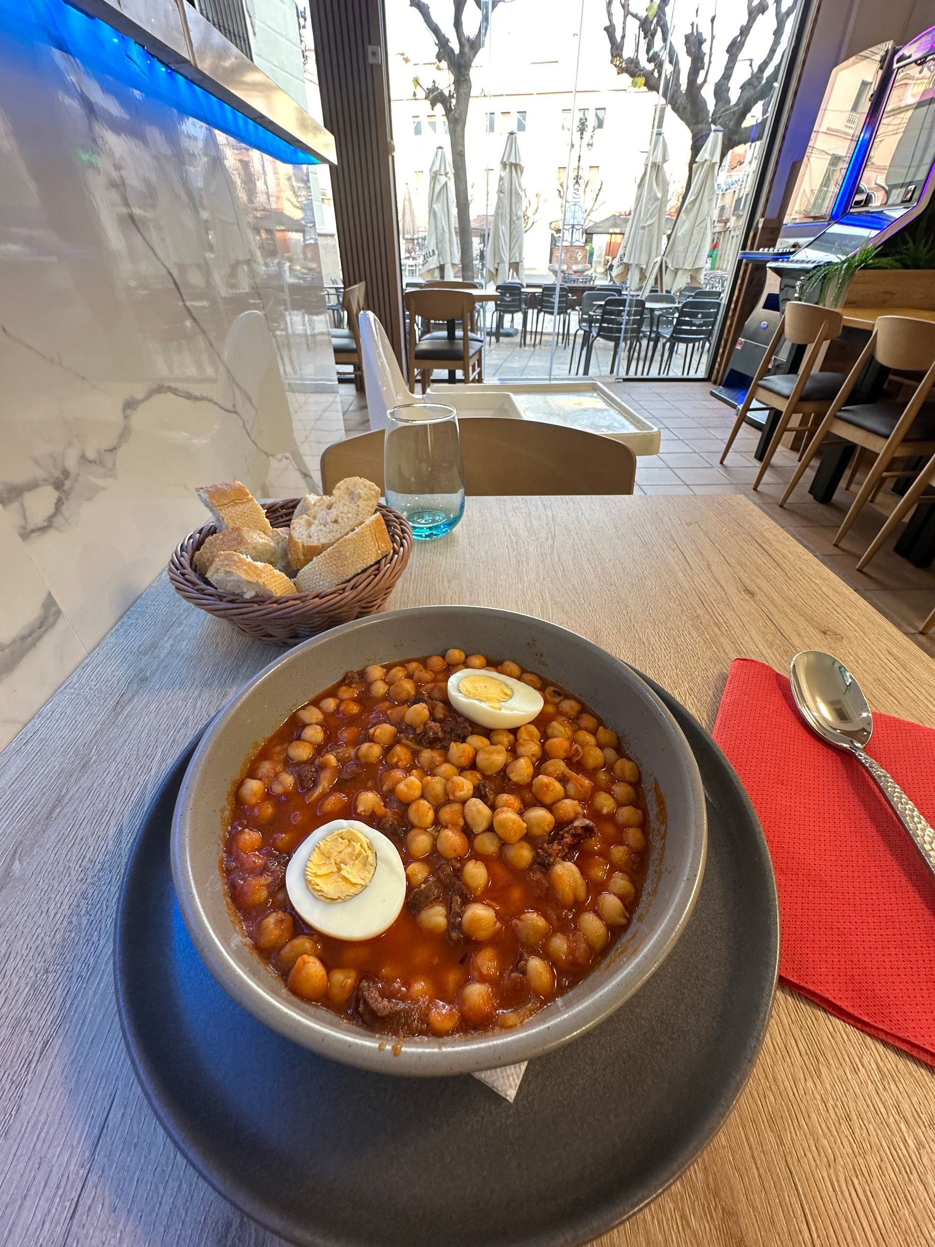 Guiso de garbanzos con huevo en cuenco gris, cesta de pan, entorno de restaurante al aire libre.