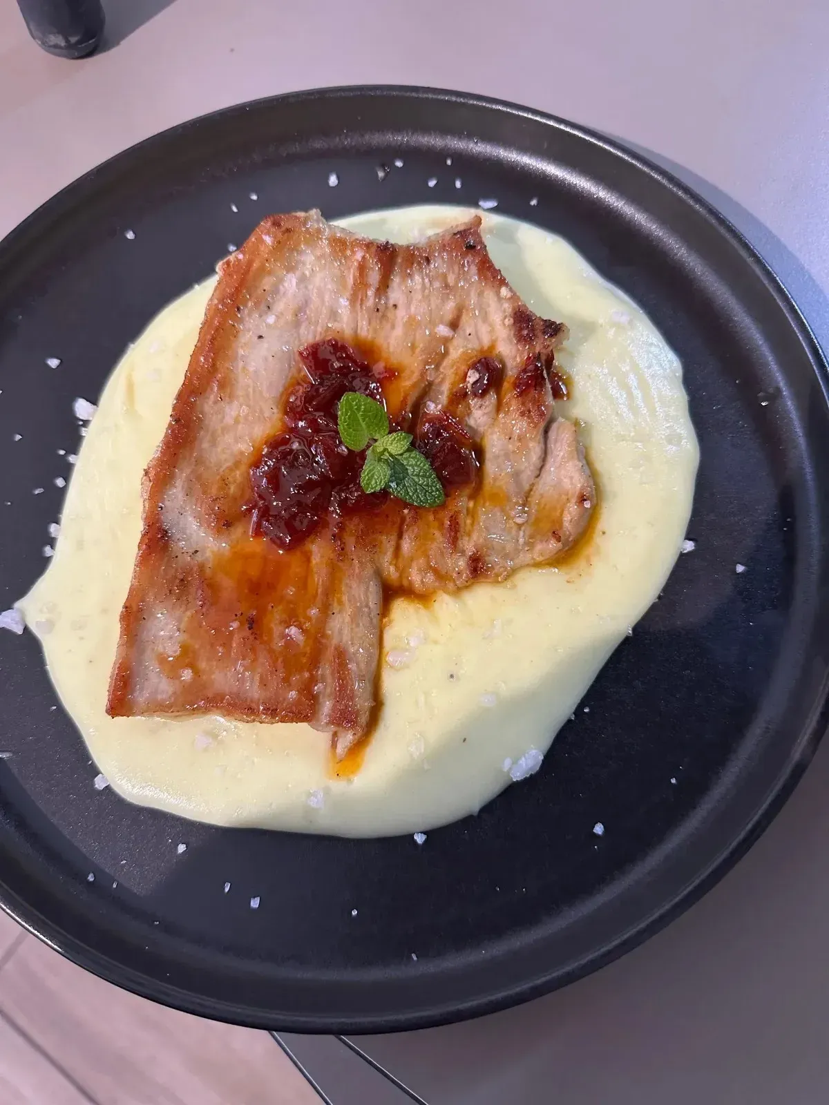 Chuleta de cerdo con salsa y guarnición de menta sobre puré de papas en un plato negro.