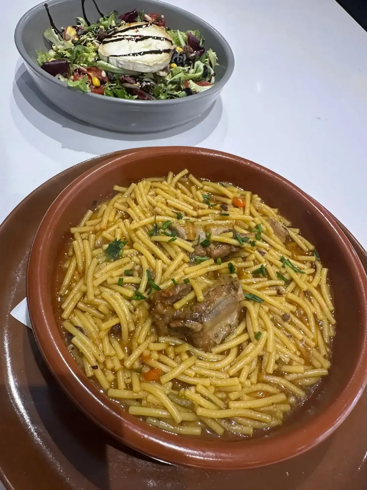 Sopa de fideos con carne en cuenco de barro, con ensalada de fondo.
