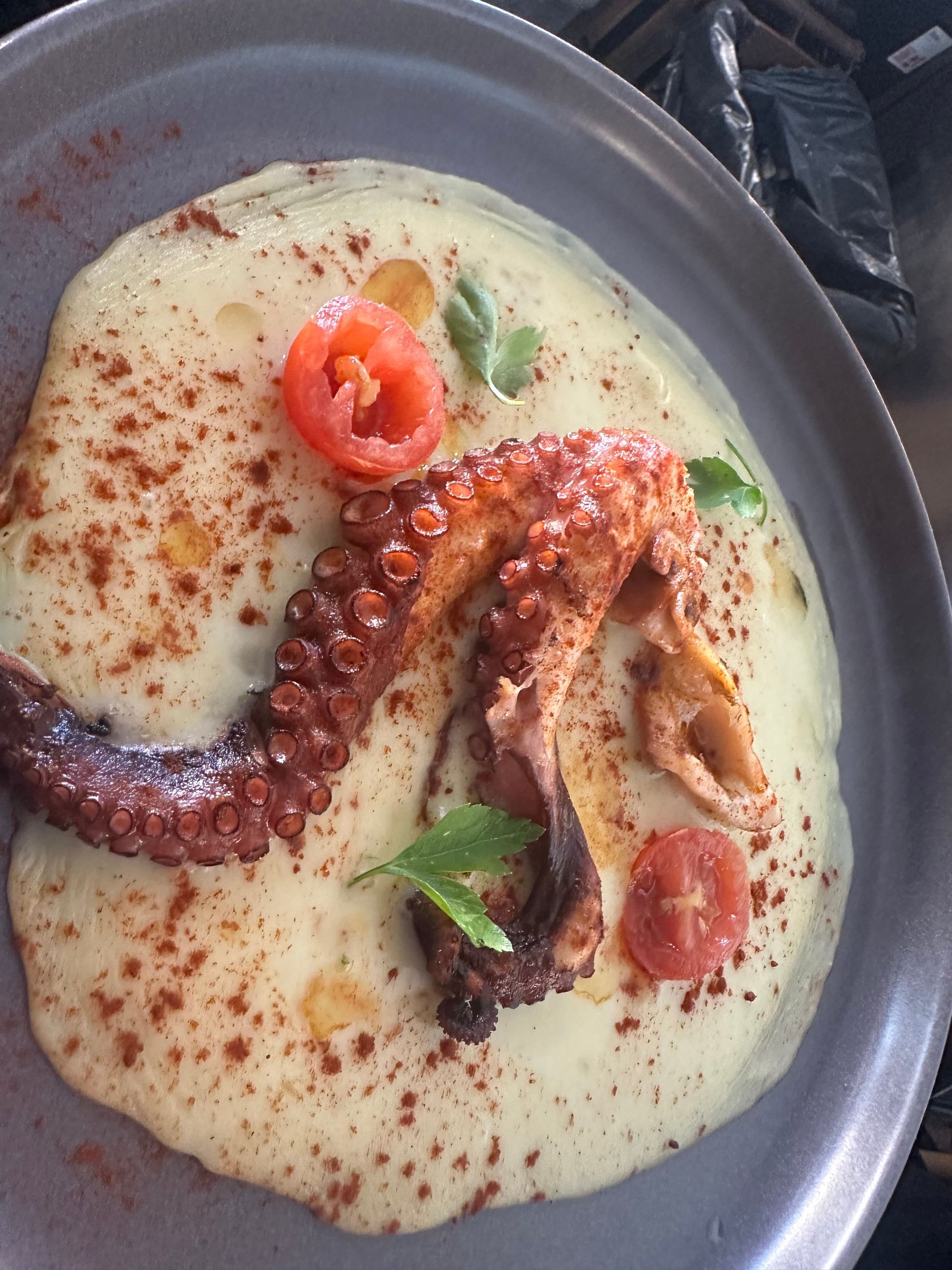 Pulpo a la plancha sobre salsa cremosa, adornado con tomate y pimentón, en plato gris.