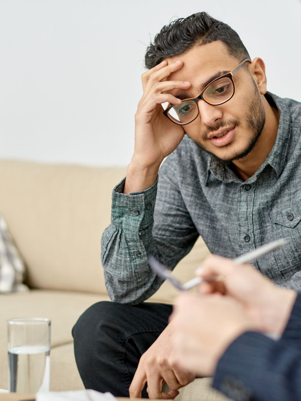 Une personne portant des lunettes est assise sur un canapé, la tête entre les mains, l'air soucieux, pendant une conversation avec une autre personne.