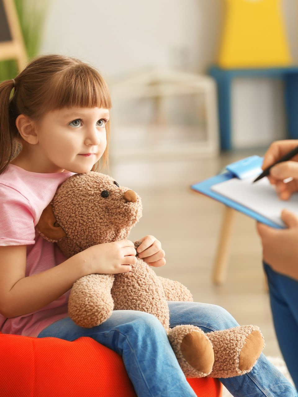 Un enfant tient un ours en peluche et regarde une personne qui prend des notes sur un bloc-notes dans une pièce bien éclairée.