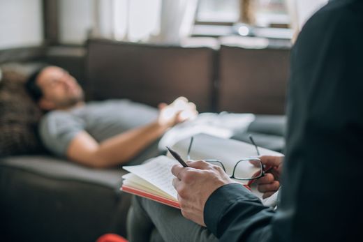 Un thérapeute professionnel, muni d'un carnet et de lunettes, écoute une personne allongée sur un divan pendant une séance.