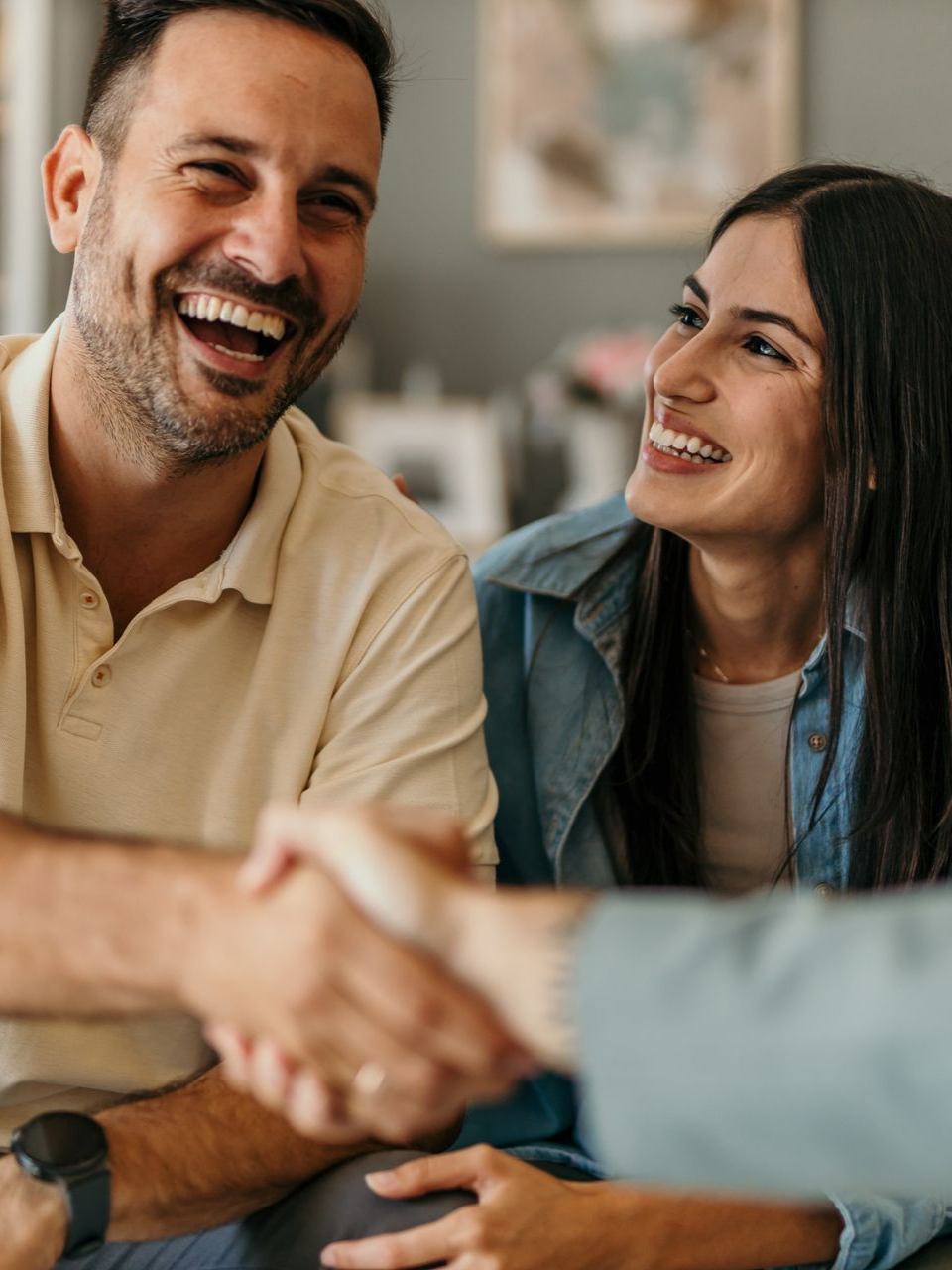 Un couple souriant serre la main à une personne hors champ dans un décor intérieur décontracté et lumineux.