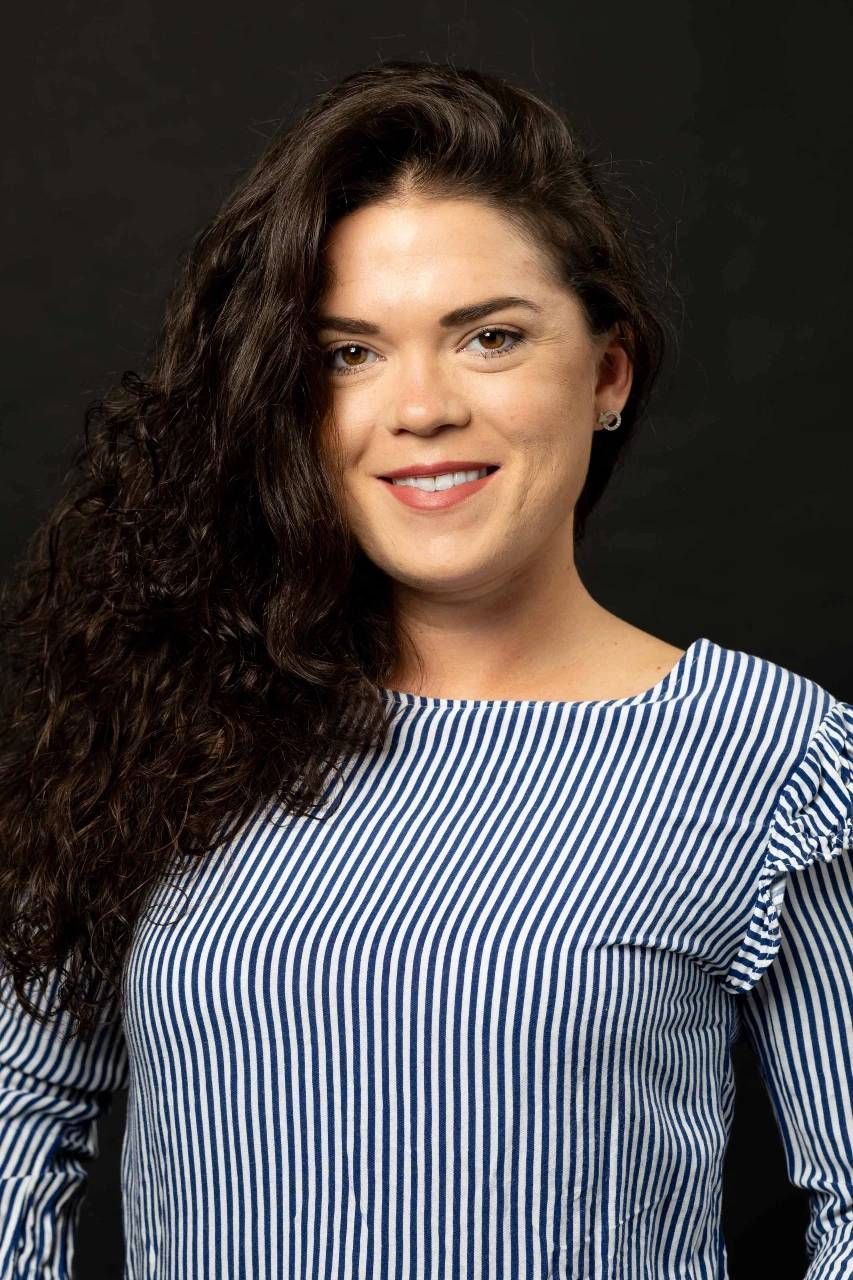 Une femme aux cheveux noirs et bouclés sourit, vêtue d'une chemise à rayures bleues et blanches.