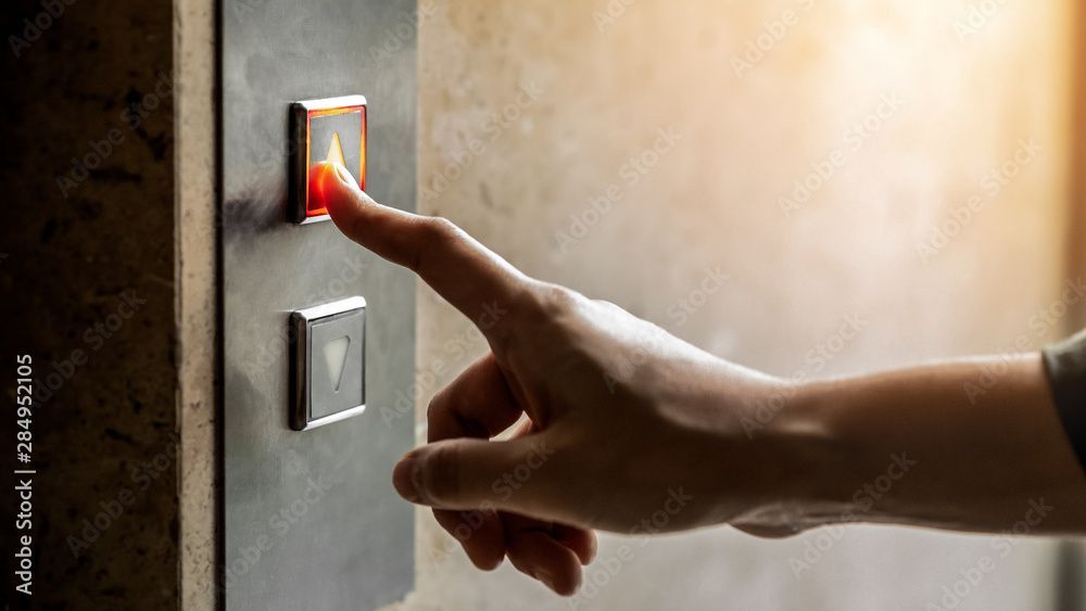 Eine Hand drückt einen beleuchteten Aufzugsknopf; beige Wand, Sonnenlicht.