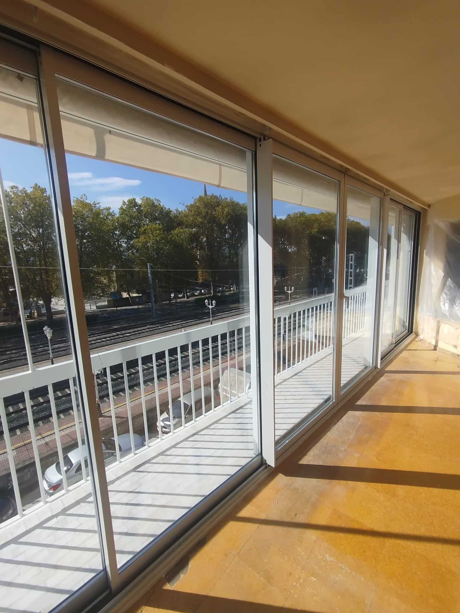 Balcón con puertas corredizas de vidrio, barandilla blanca y vista a los árboles y al cielo.