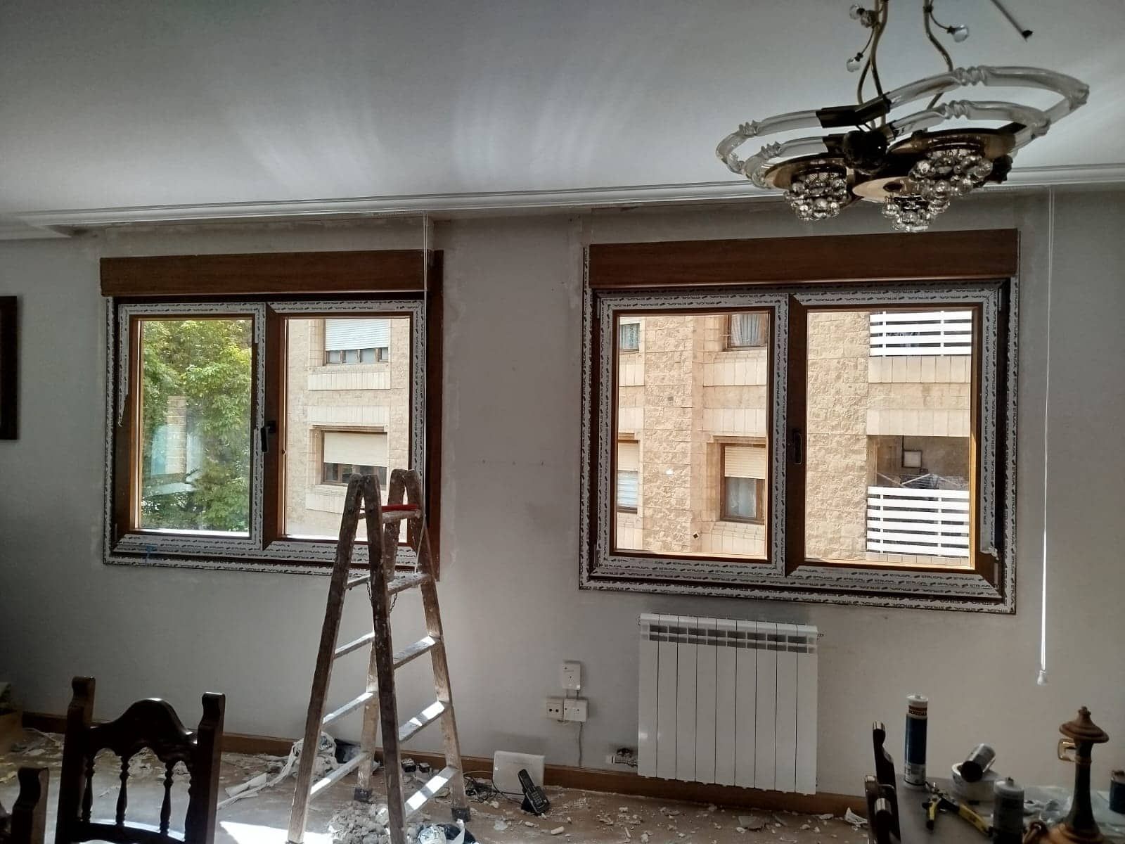 Dos ventanas en una habitación con escalera, radiador y lámpara de araña. Se ven escombros de construcción.