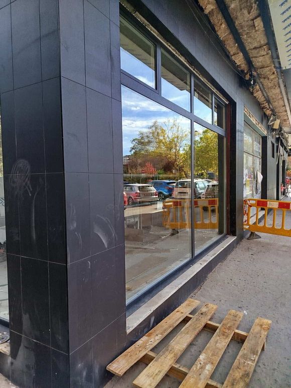 Fachada exterior de tienda con gran ventanal que refleja una escena callejera, pilar de azulejos negros y palé de madera en primer plano.