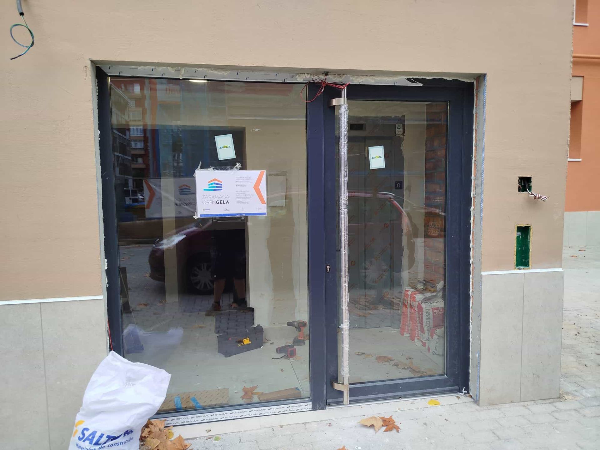 Exterior de un local comercial con puerta y ventana de cristal. Se ven materiales de construcción cerca de la entrada.