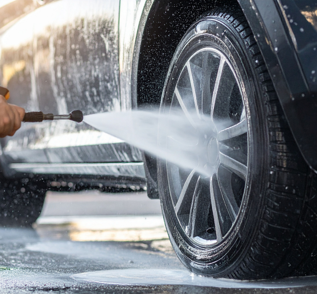 Une personne lave le pneu d'une voiture noire avec un pulvérisateur d'eau, créant un jet d'eau.
