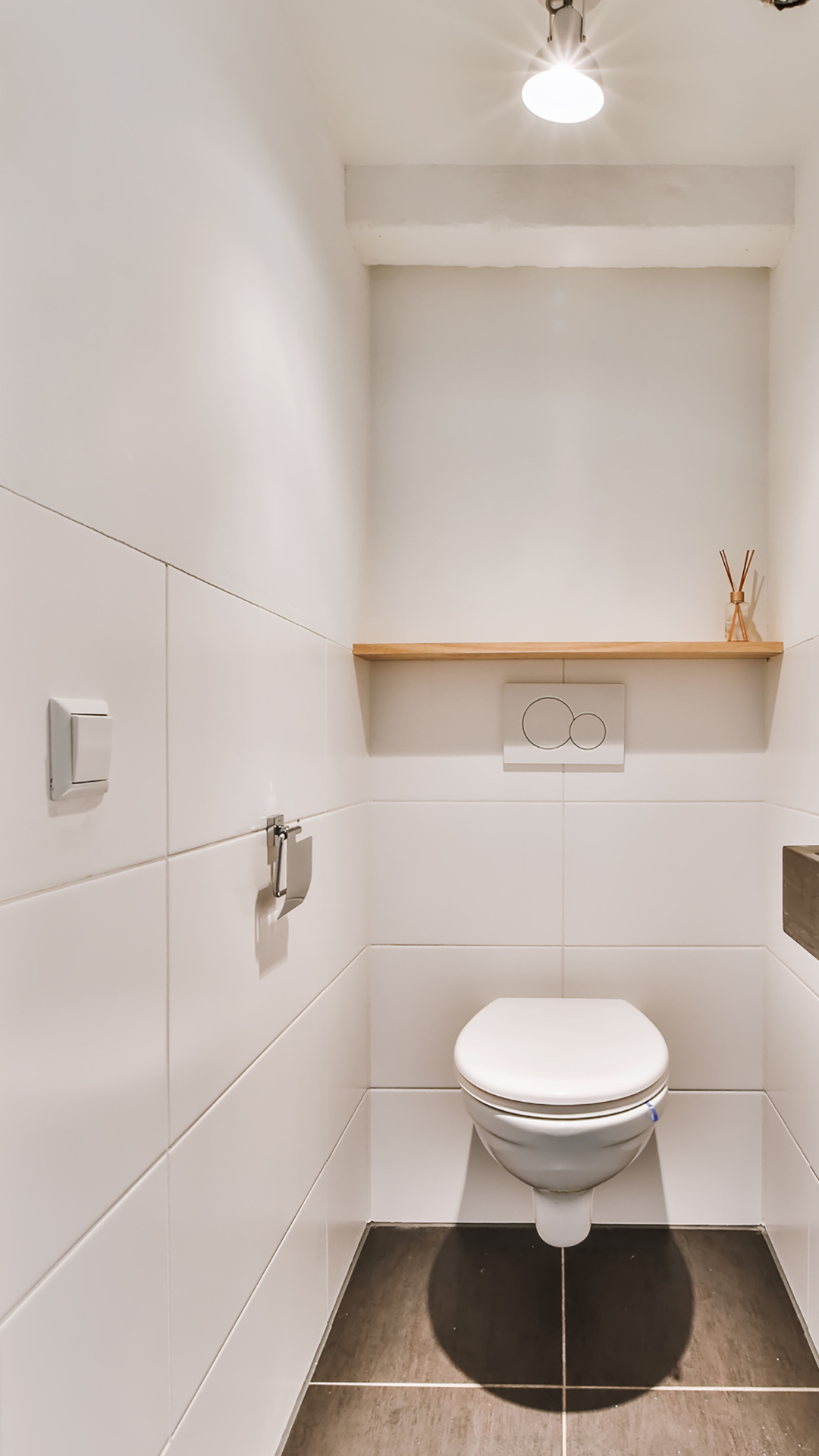 Salle de bain étroite et moderne avec murs carrelés blancs, toilettes et une petite étagère en bois.