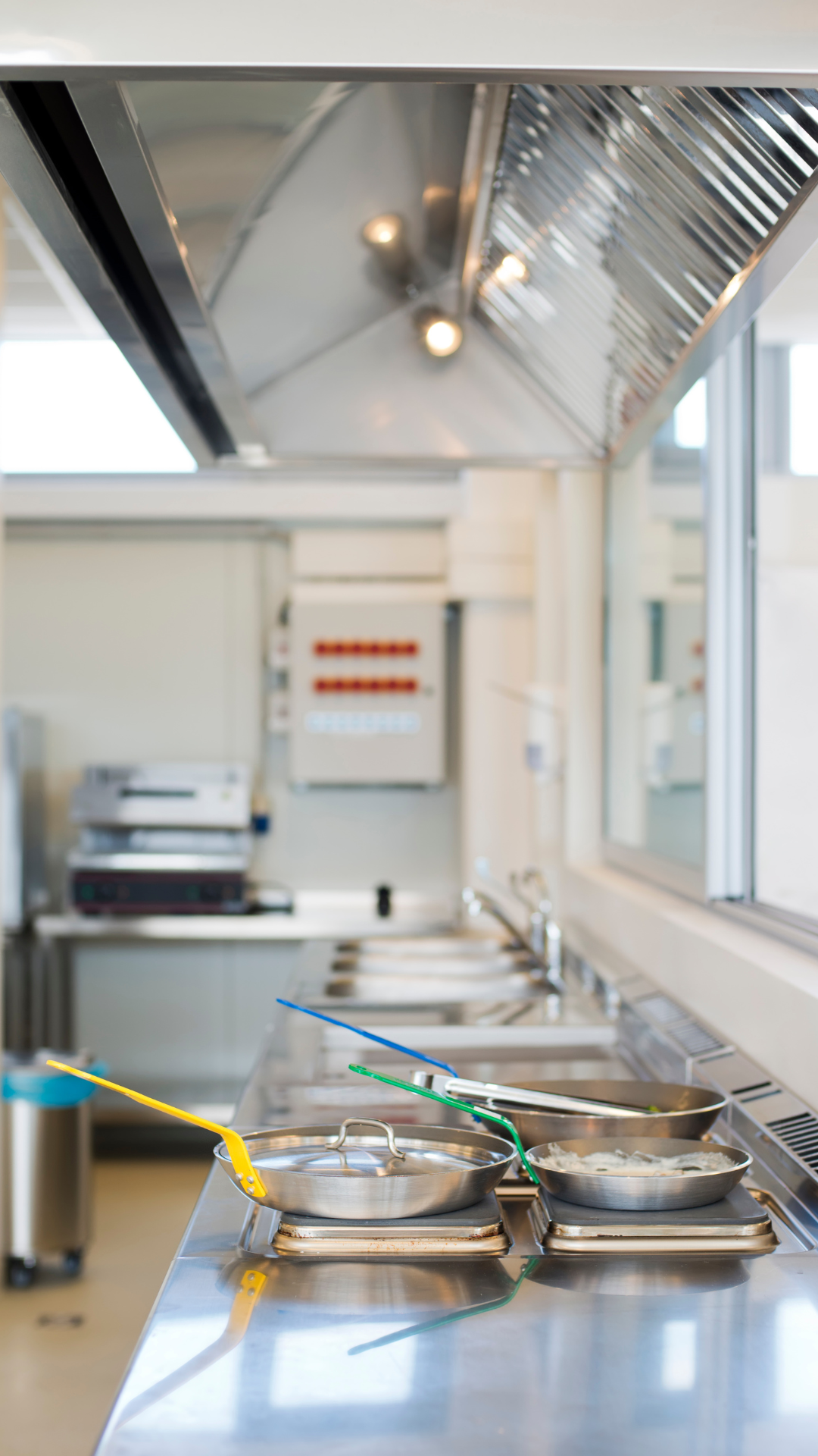 Cuisine commerciale avec comptoirs en acier inoxydable, cuisinière, hotte aspirante et ustensiles de cuisine.