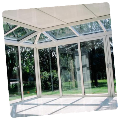 Véranda avec murs et plafond en verre, cadre blanc, arbres visibles à l'extérieur.