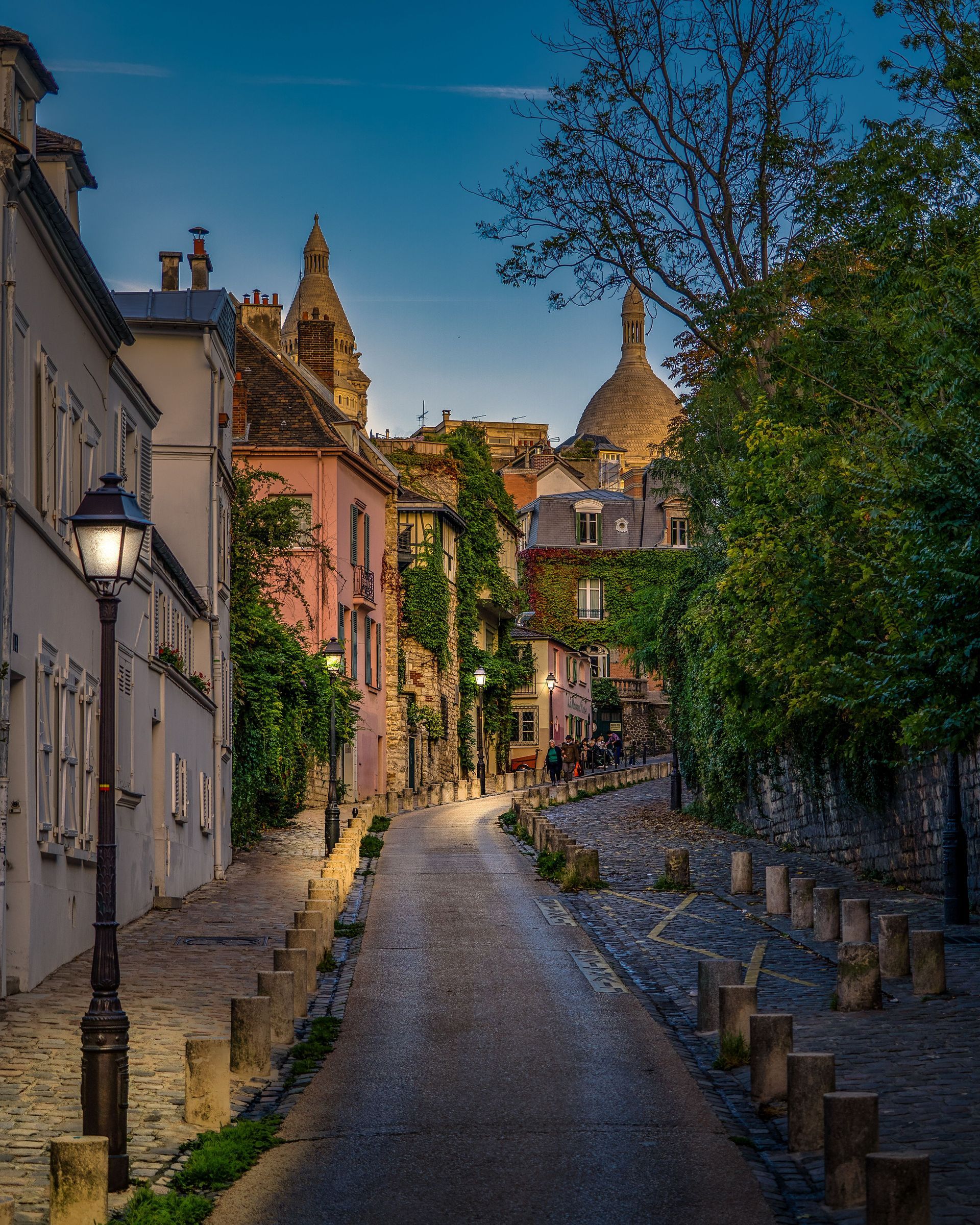 Narrow cobblestone street in a sunlit village, lined with ivy-covered houses and church domes at dusk.