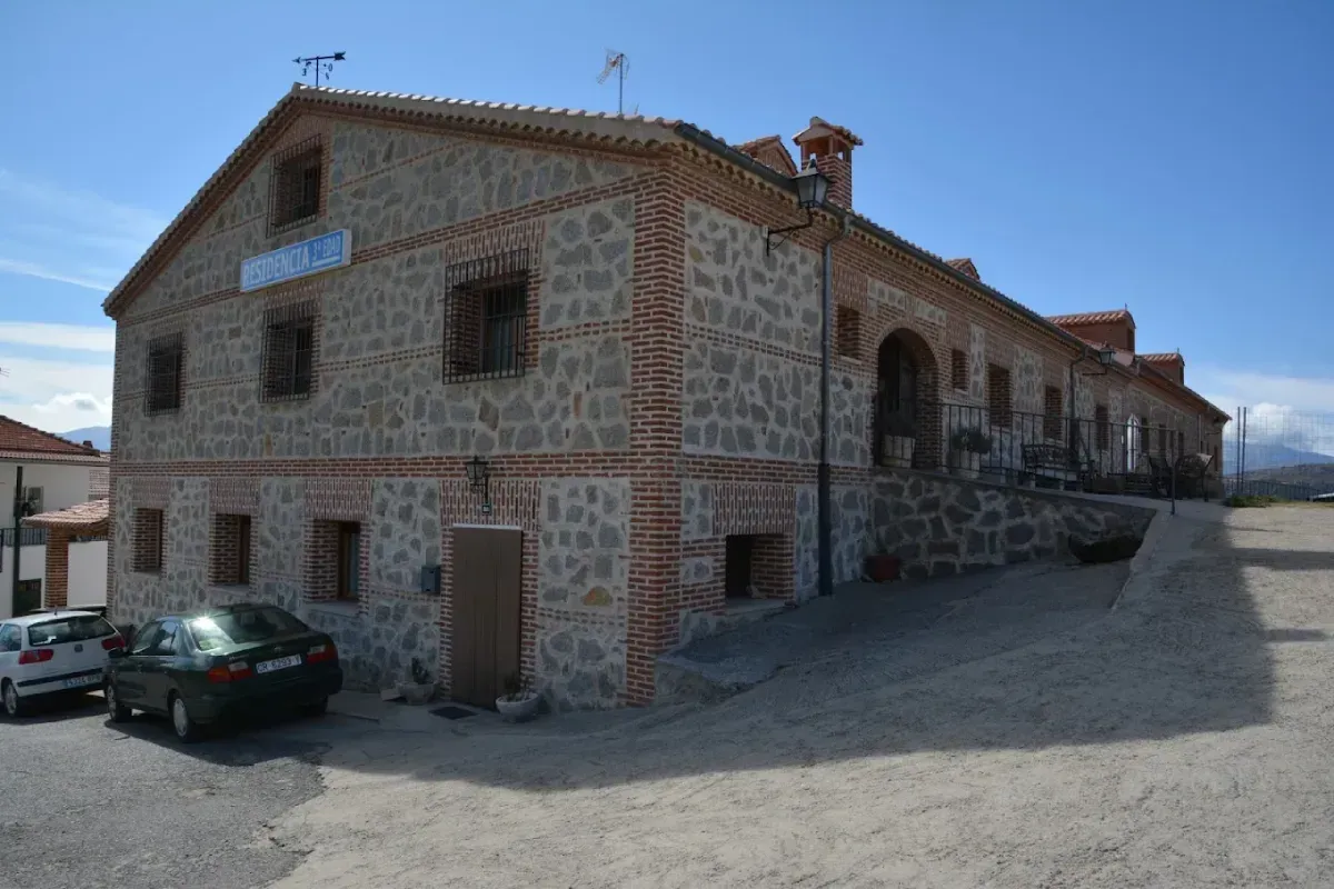 Edificio de piedra con un letrero azul, un coche verde aparcado y una calle empedrada bajo un cielo despejado.