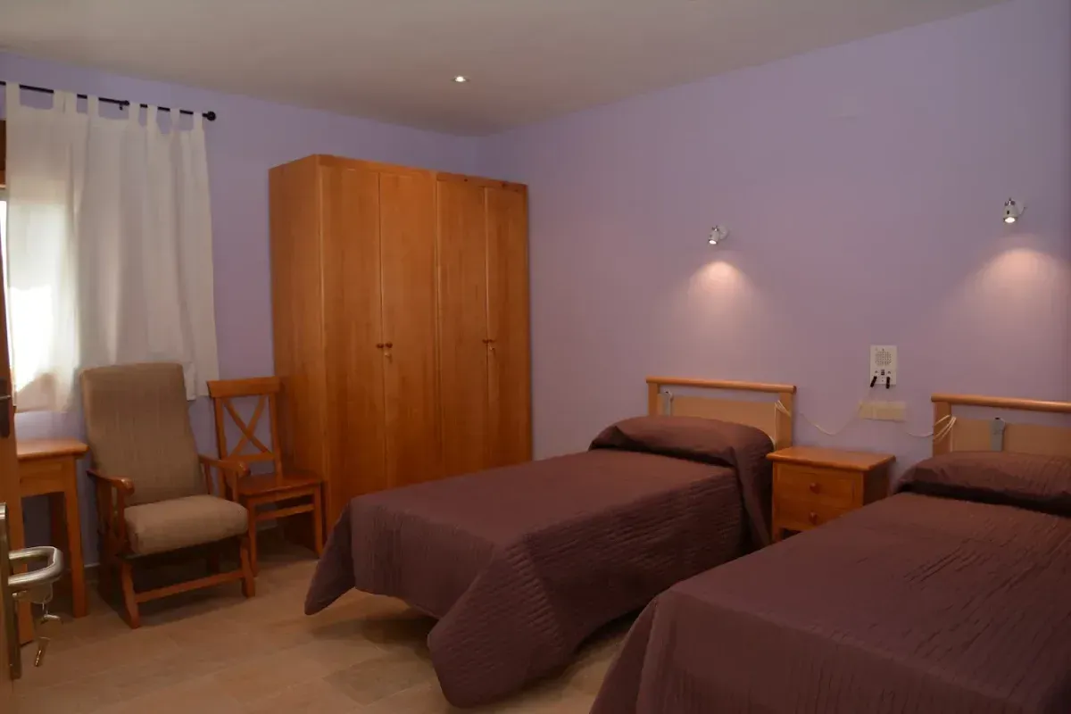 Dos camas individuales en una habitación de color lavanda con armario, sillas y ventana con cortinas.