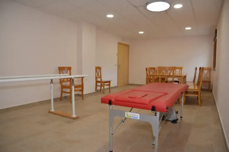 Habitación vacía con una camilla de masaje roja, sillas de madera y paredes beige bajo luces de techo.
