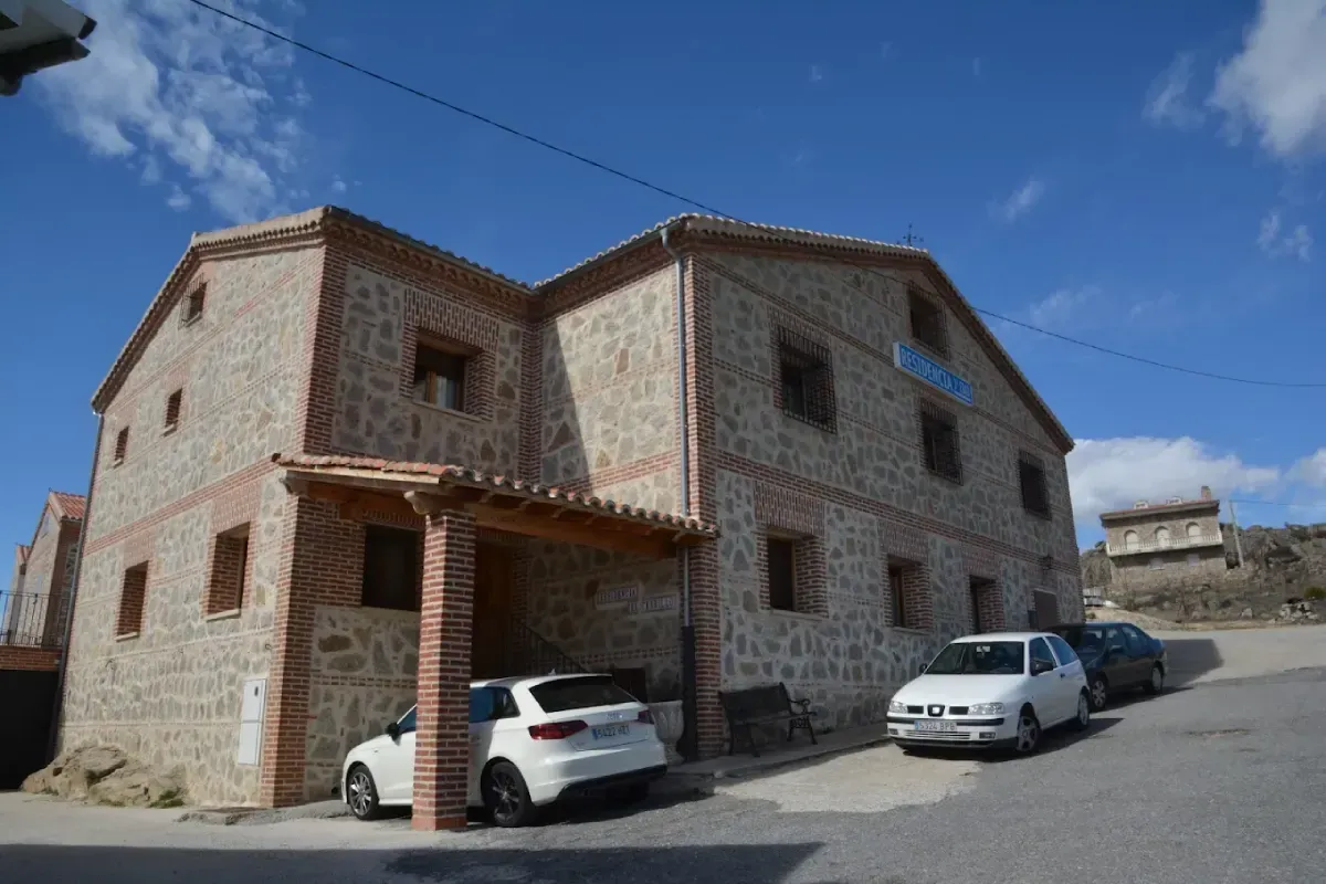 Casa de piedra en una calle en pendiente con dos coches aparcados bajo un cielo azul.