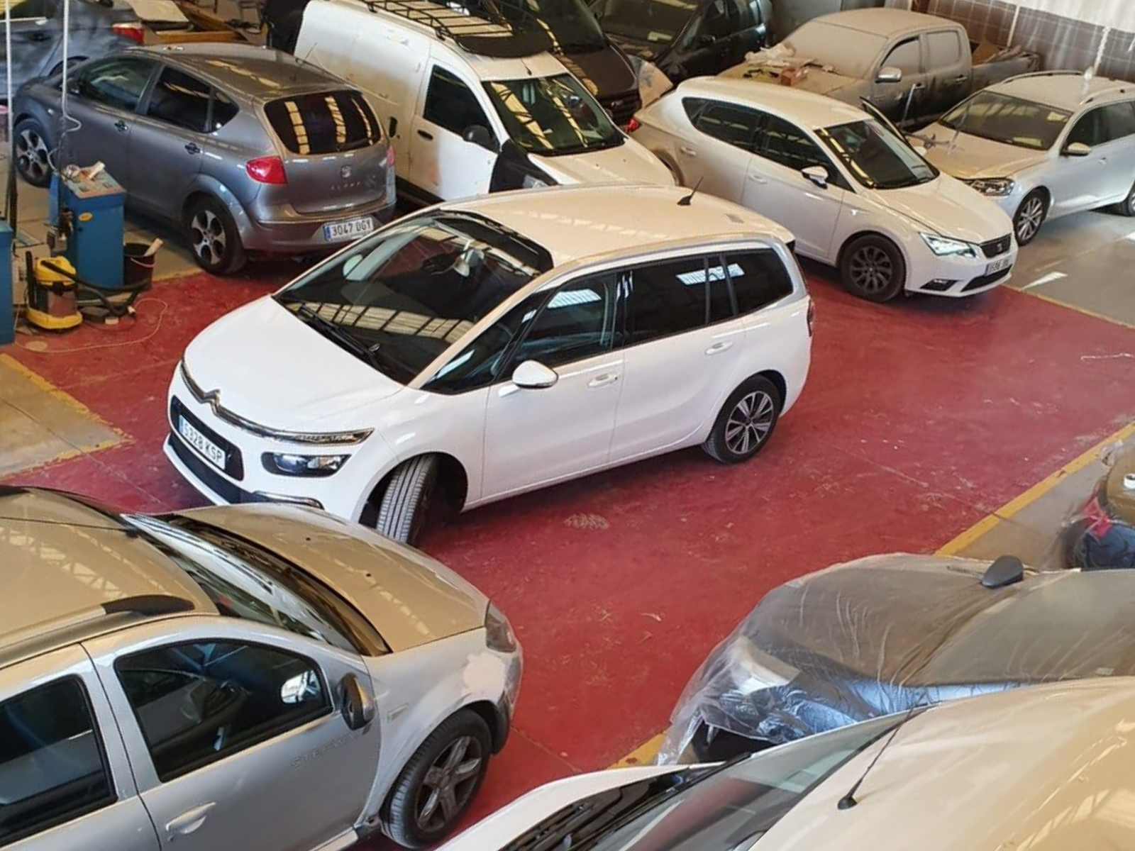 Minivan blanca rodeada de varios autos estacionados, dentro de una estructura tipo garaje con piso rojo.