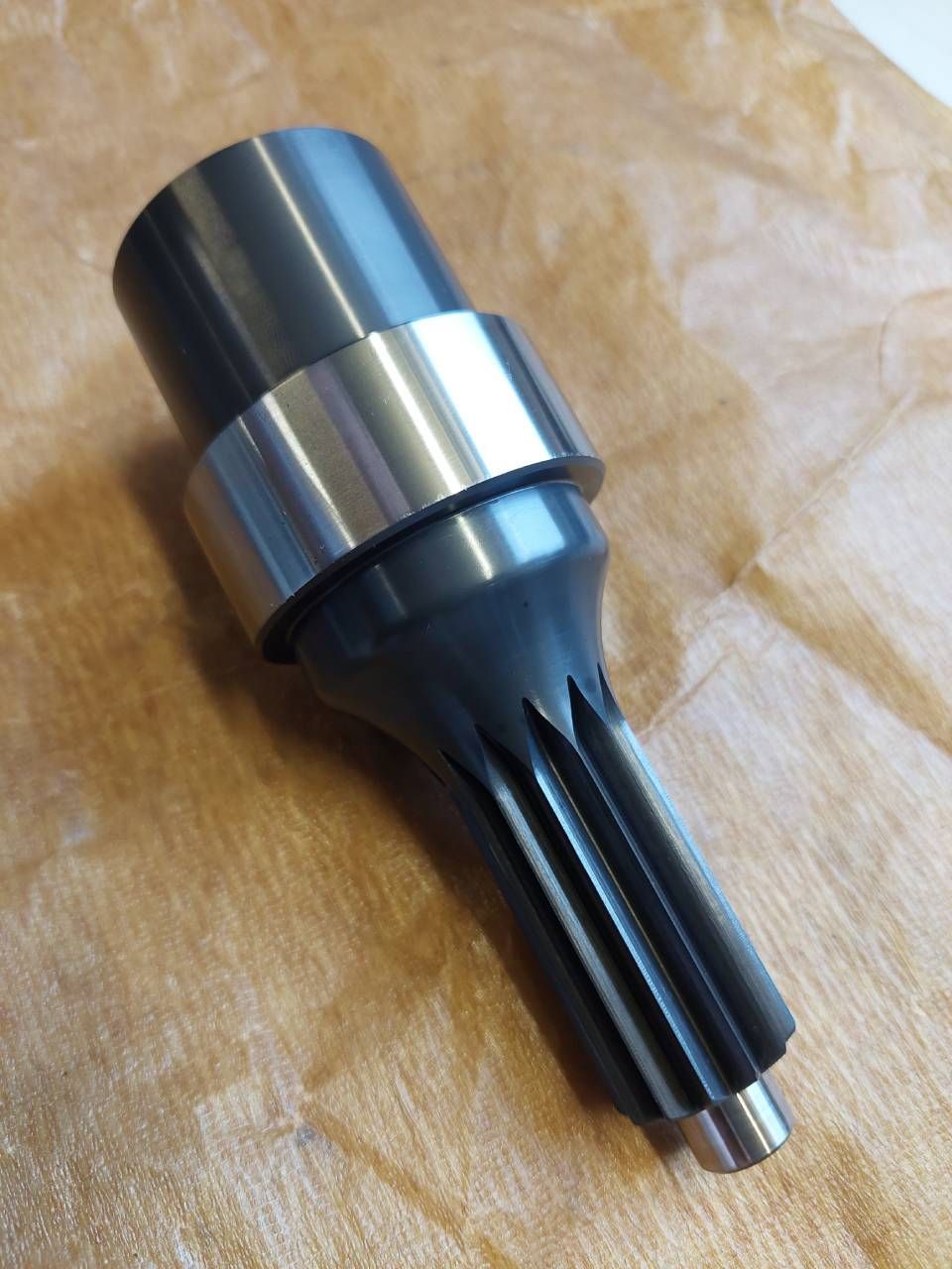 Metallwelle mit Zahnradverzahnung, zylindrischen Abschnitten und einem Lagerring auf braunem Papier.