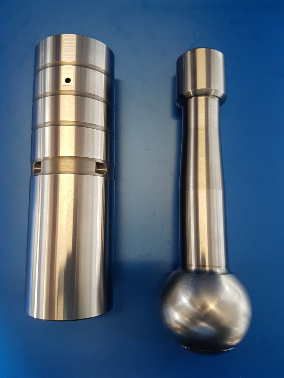 Zwei bearbeitete Metallkomponenten auf einer blauen Oberfläche; ein zylindrisches Teil und ein Teil mit einem kugelförmigen Ende.