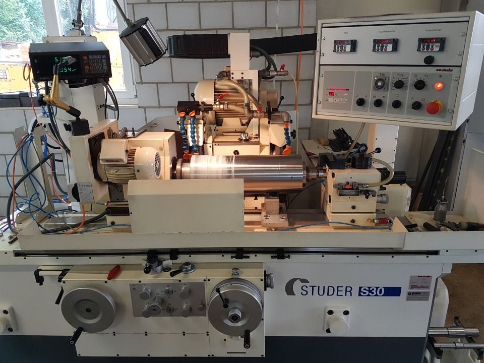 Eine Rundschleifmaschine in einer Werkstatt mit der Aufschrift „STUDER S-30“ hat einen eingespannten Metallzylinder.