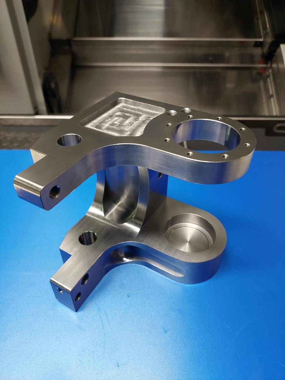 Bearbeitetes Metallteil mit mehreren Armen, Löchern und einer gemusterten Oberseite, das auf einer blauen Oberfläche ruht.