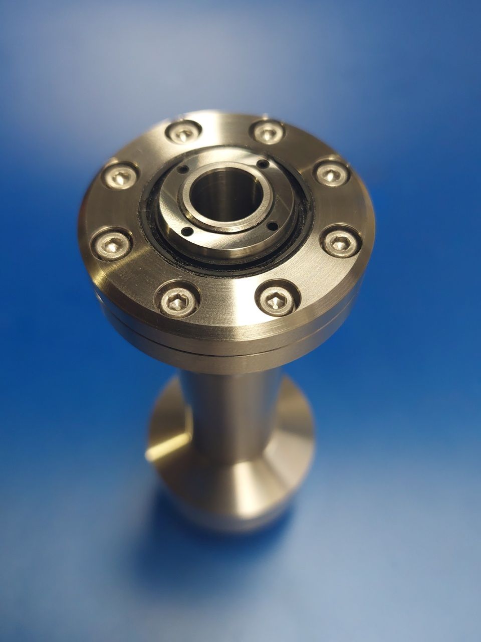 Industrielles Bauteil aus Edelstahl mit runder Oberseite und Befestigungslöchern auf blauem Hintergrund.