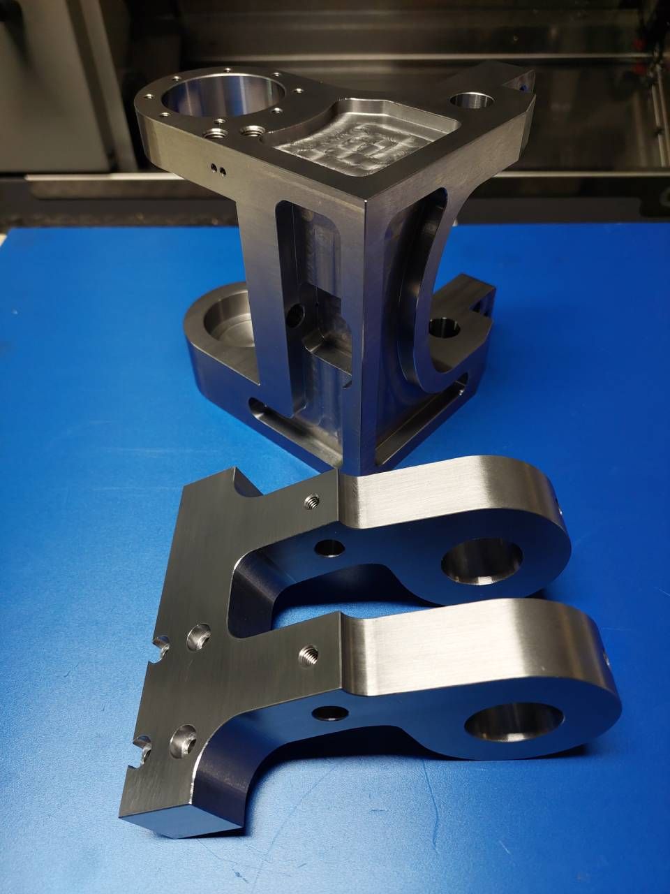 Drei bearbeitete Metallteile auf blauem Untergrund. Ein grösseres, zwei kleinere, alle mit Löchern und geometrischen Formen.