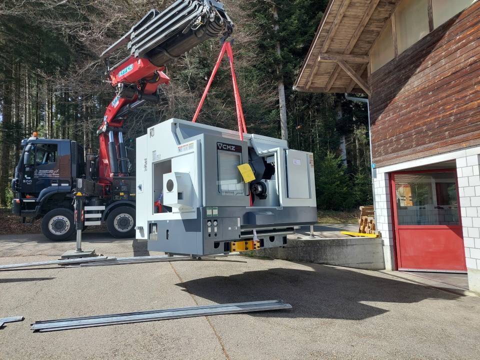 Eine grosse CNC-Maschine wird in der Nähe eines Gebäudes mit einem Kran angehoben.