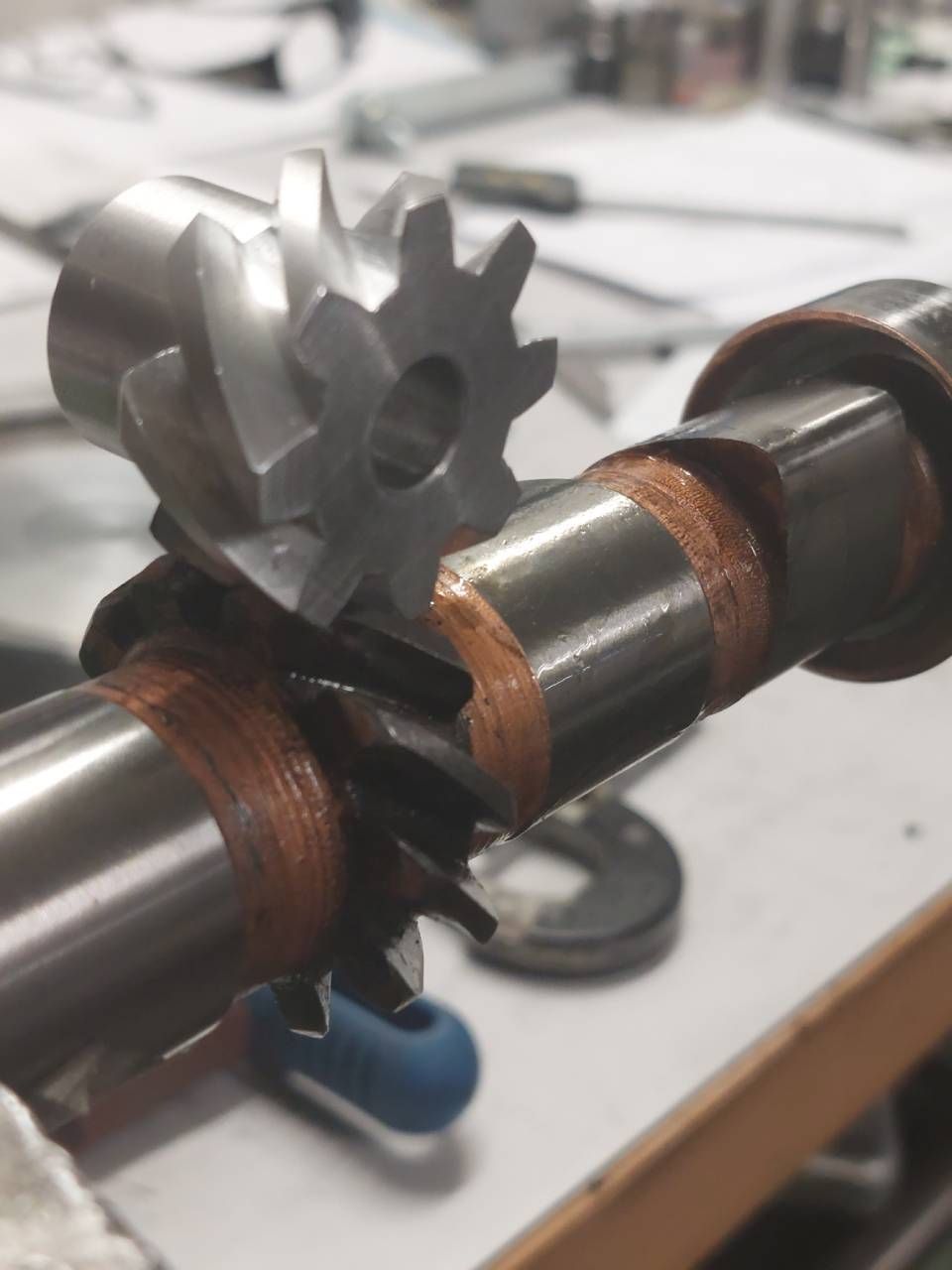 Zwei Zahnräder greifen auf einer Metallwelle ineinander, vermutlich Teil einer Maschine oder eines Motors. Die Welle ist mit braunem Papier umwickelt.