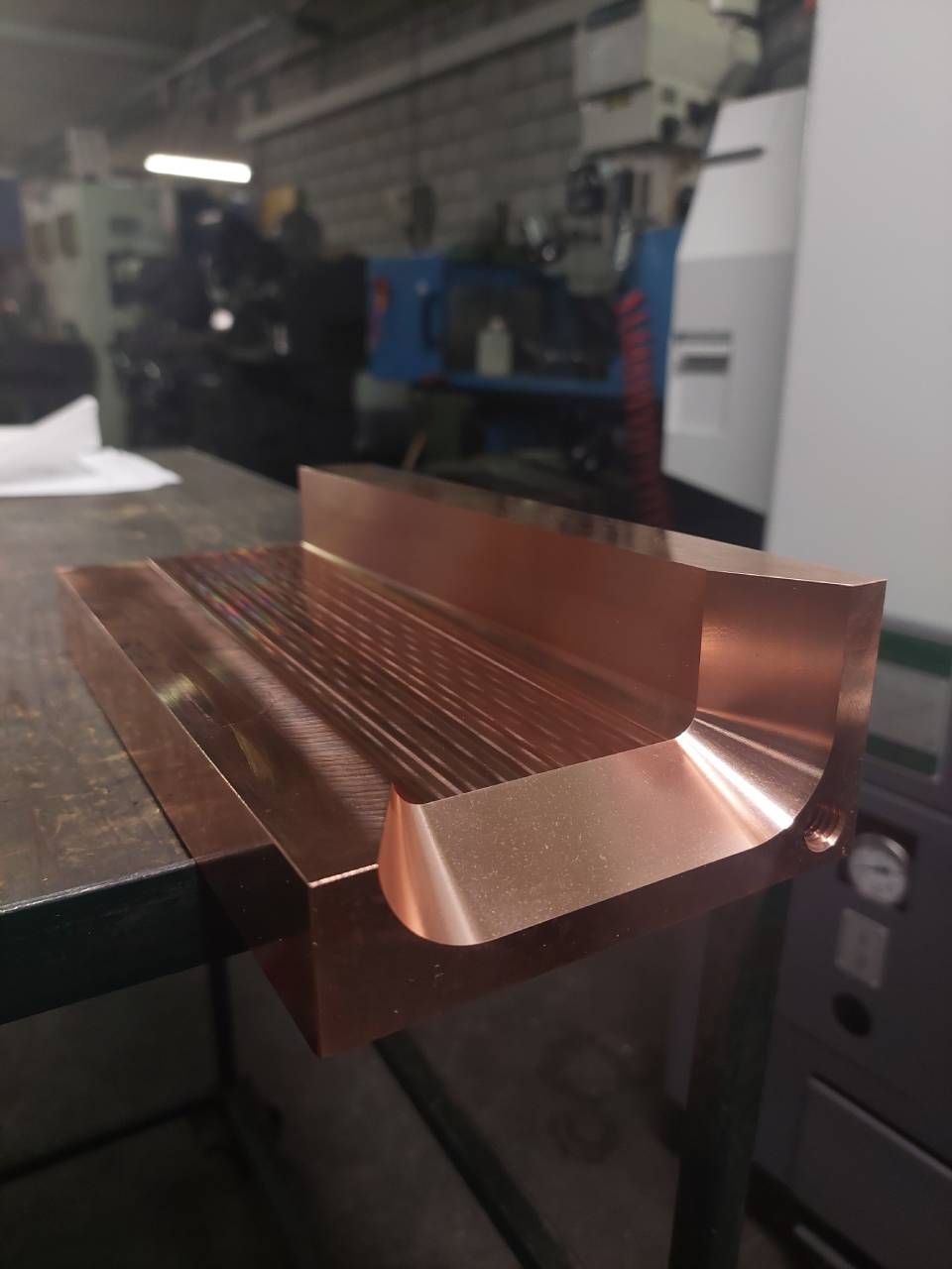 Kupferfarbenes, bearbeitetes Metallteil auf einer Werkbank in einer Werkstatt. Gerillte Innenseite, abgeschrägte Kanten und Maschinen im Hintergrund.
