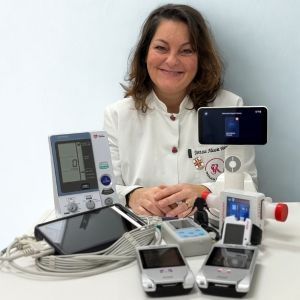 Una donna mora con i capelli che appoggiano alle spalle in camice sorride, circondata da dispositivi medici. Stanza bianca.