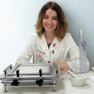 Una giovane donna con capelli biondo cenere dino alle spalle in camice sorride mentre è seduta al banco del laboratorio con l'attrezzatura.