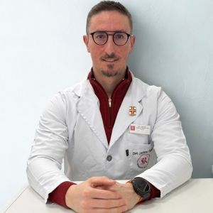 Uomo moro con baffi, pizzetto e occhiali in camice bianco , seduto, braccia incrociate.