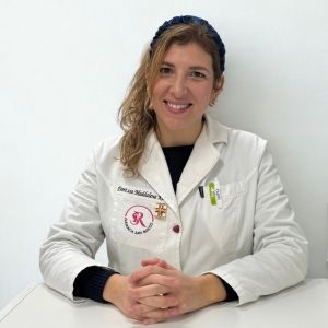Donna sorridente, capelli biondi alle spalle con camice bianco, seduta, con uno stetoscopio in tasca.