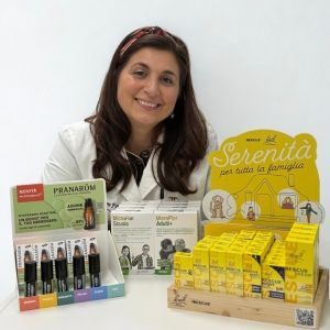 Giovane donna con capelli lunghi castano scuro sorride, in piedi dietro gli espositori di prodotti con oli essenziali e integratori. Sfondo bianco.