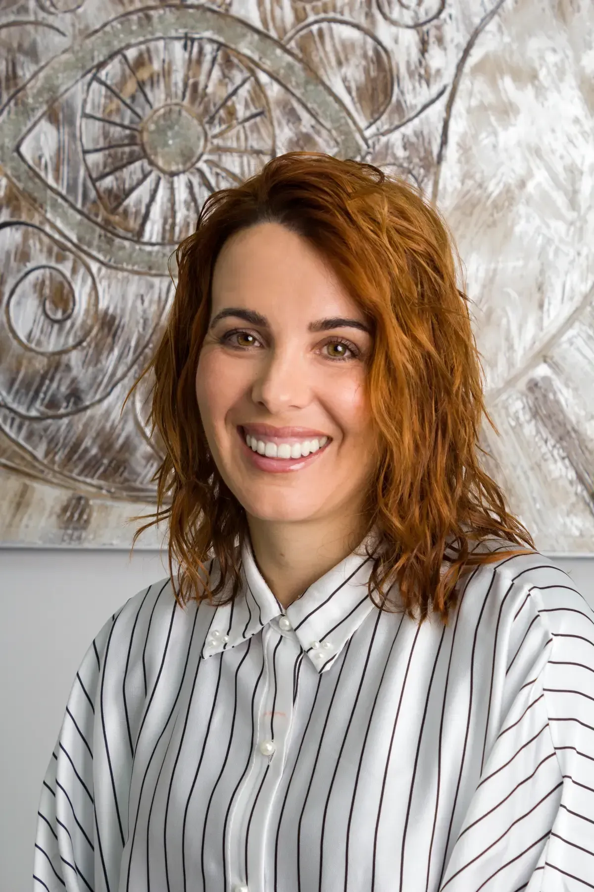Una persona sonriente con cabello rojo ondulado que viste una camisa blanca con finas rayas negras