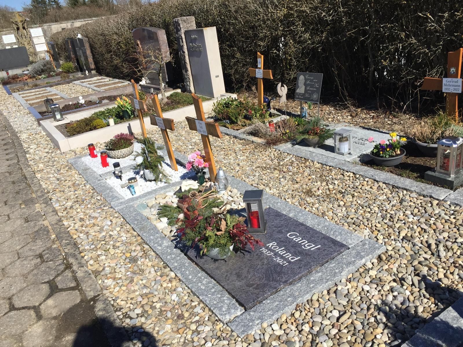 Grabstätten auf einem Friedhof. Einige sind mit Kreuzen, Grabsteinen und Blumen geschmückt. Der Boden ist mit Kies bedeckt.